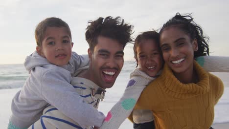 Portrait-of-happy-hispanic-parents-carrying-piggyback-children-on-beach