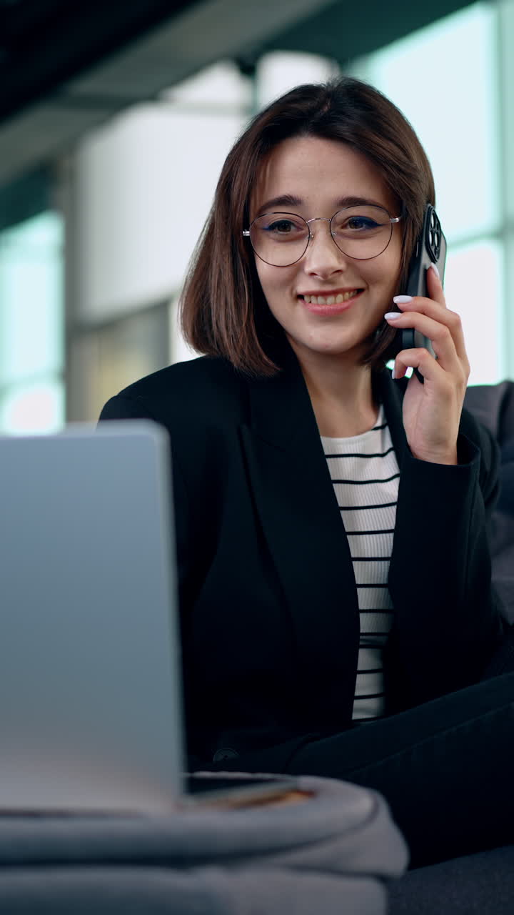 Cute smiling brunette girl holds the phone at her ear. Lady looks at her laptop and feels excited. Vertical video.