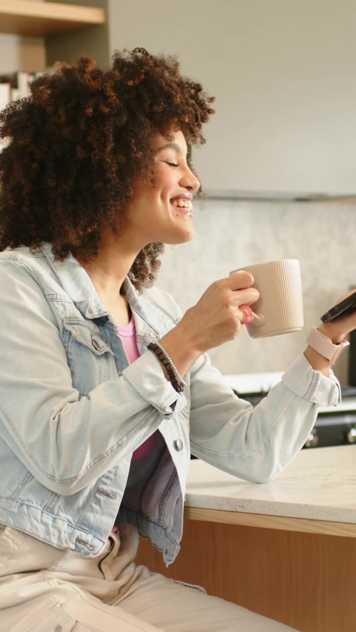 Vertical video: Checking woman in denim jacket reading text at home counter, holding phone and mug
