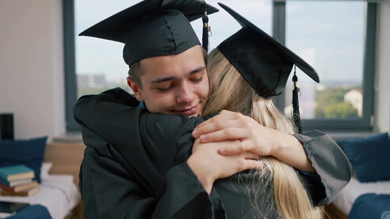 Laureati che si incontrano e si abbracciano vicino alla finestra in un dormitorio, festeggiando con toghe, cappelli da laurea e mani giunte.