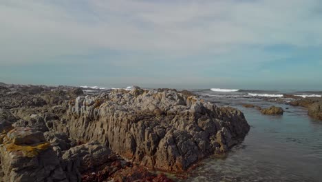 Drone-flying-low-over-shore-front-with-boulders-and-rocks-revealing-open-ocean-waters-with-bamboo-and-rolling-waves