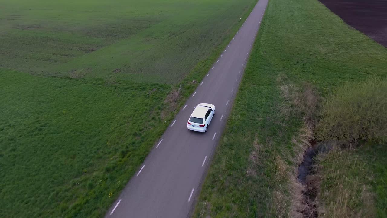 Ripresa aerea con drone e prospettiva a volo d'uccello di un'auto bianca che percorre una stretta strada di campagna nella Scania, Svezia meridionale.