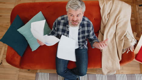 from above shot stressed angry mature man reviewing charts graphs throwing papers documents on sofa