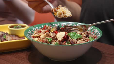 two people use spoon and fork to eat a colorful thai beef, garlic, and chili rice meal in a casual bangkok restaurant with warm lighting and close-up camera angles