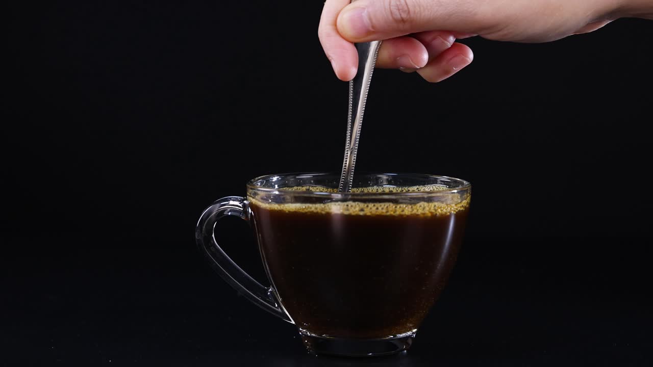 A hand stirs coffee in a glass mug against a dark backdrop, highlighting the swirling motion and rich color