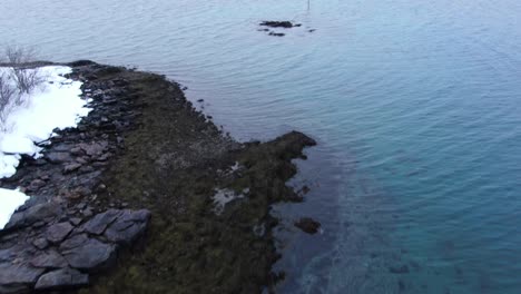 Vista-De-Drones-En-Tromso-Volando-Hacia-El-Mar-Desde-Una-Zona-Nevada-Con-Montañas-Llenas-De-Nieve-En-Invierno-En-Noruega