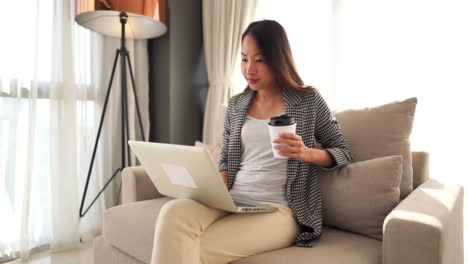 Eine-Hübsche-Asiatin,-Die-Auf-Einem-Gepolsterten-Stuhl-Sitzt-Und-Eine-Kaffeetasse-Hält,-Während-Sie-Auf-Ihrem-Laptop-Tippt