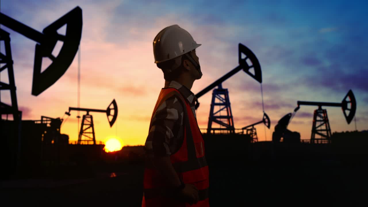 vista lateral de un ingeniero masculino asiático con casco de seguridad mirando a su alrededor mientras está de pie con los brazos akimbo frente a las bombas de petróleo, durante la puesta o salida del sol