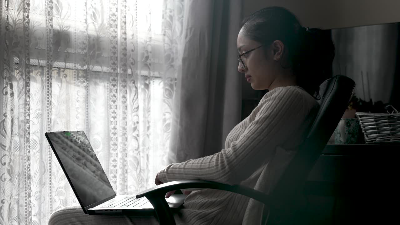 Asian British Housewife Sitting Near Window working on a laptop placed on their lap, showcasing the integration of digital media into home environments
