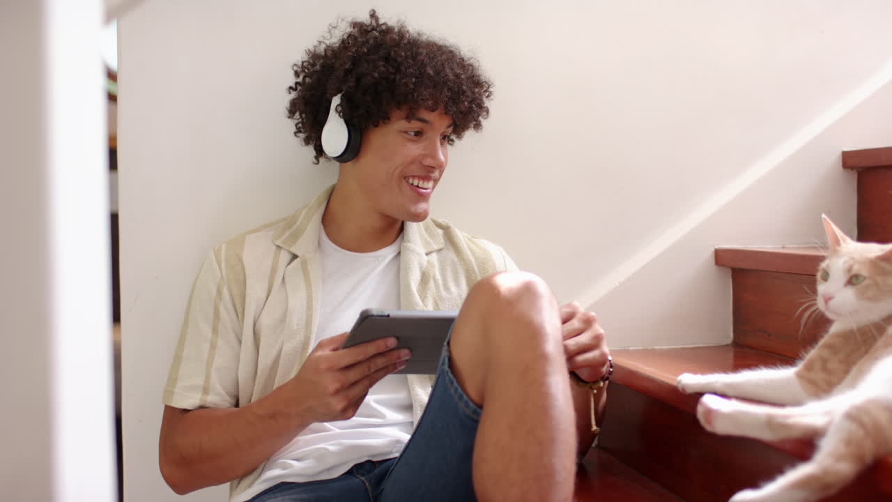 Ascoltare musica con le cuffie, uomo che gioca sul tablet e sorride al gatto, a casa