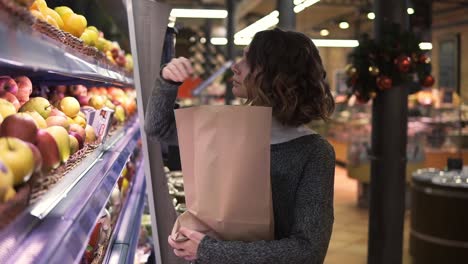 Cute-girl-buys-fresh-red-apples-in-the-market.-Beautiful-young-woman-stands-in-front-the-shelf-and-puts-the-apples-to-a-brown-paper-bag,-she-is-pleased-with-the-choice.-Slow-motion