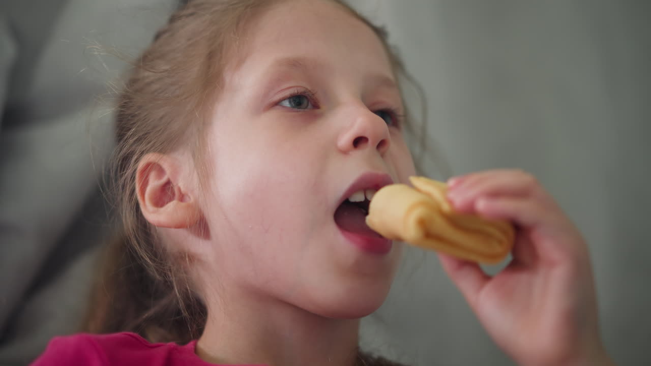 Close up of cute kid in pink top shifting snack to left hand before taking bite with focused expression indoors showing natural childhood behavior