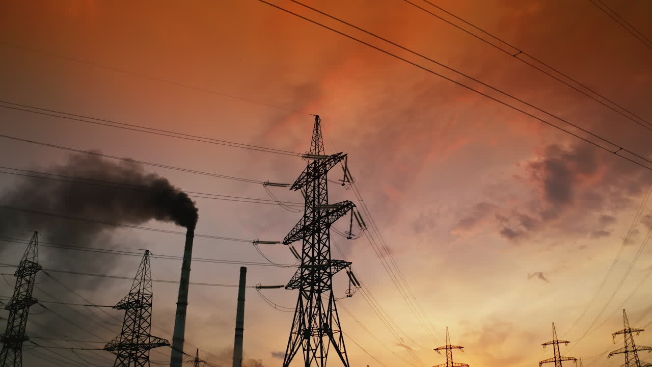 Electric towers near factory chimney with dark smoke. Transmission lines on background of harmful fumes from industry releasing from pipes at sunset. Environment pollution.