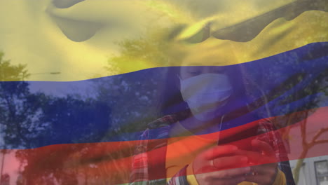 bandera colombiana ondeando contra una mujer con máscara facial usando un teléfono inteligente