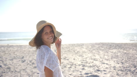 young biracial woman smiles on a sunny beach, wearing a white dress and a straw hat, with copy space