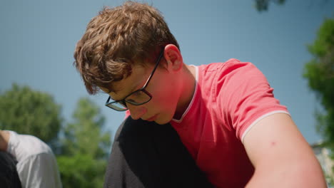 un niño con una camisa roja se arrodilla trabajando intensamente en algo, ajustando sus gafas mientras se concentra, cerca, alguien está sentado cerca
