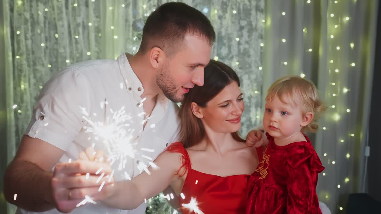Happy family, mother, father and daughter, wearing elegant clothes, holding sparklers and celebrating Christmas or New Year's Eve near a decorated Christmas tree with fairy lights