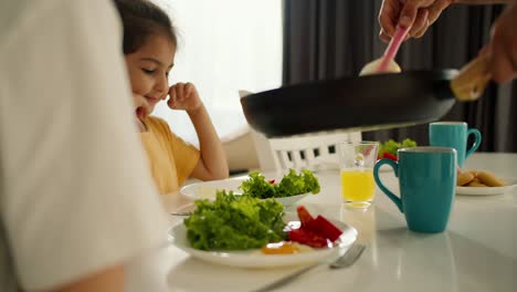 A-man-prepared-a-morning-breakfast-and-puts-scrambled-eggs-on-plates-for-his-little-daughter,-a-brunette-girl-in-a-yellow-dress,-and-for-his-wife,-a-girl-in-a-white-T-shirt,-during-the-morning-breakfast-with-salad-at-the-table-in-the-kitchen