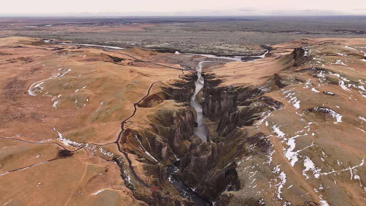 Epic aerial view of Fjaðrárgljúfur Canyon in Iceland, showcasing steep, rugged cliffs, a winding river, and expansive golden landscapes stretching toward the horizon.