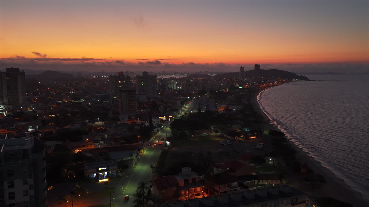Aerial view of Penha, State of Santa Catarina, Brazil at sunset showing illuminated cityscape, beach, and ocean with orange sky, creating a mesmerizing coastal urban landscape