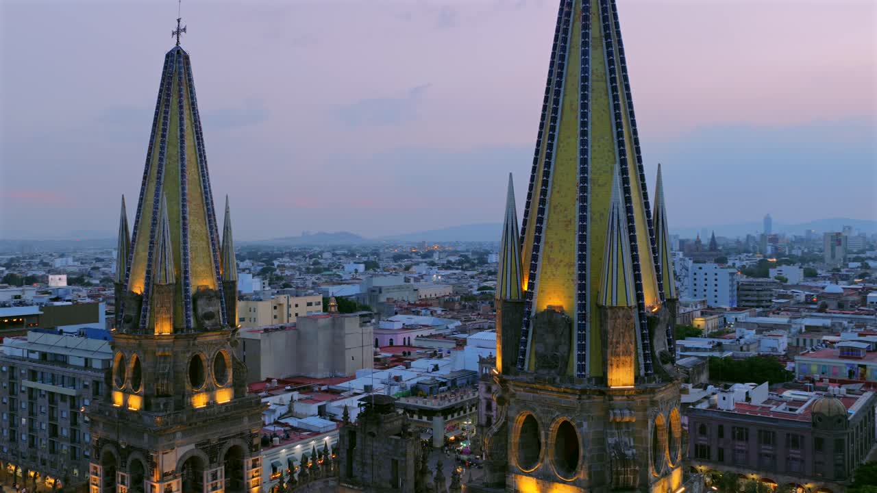 Illuminated twin spires of the Guadalajara Cathedral rise over the city center skyline at dusk, close-up aerial view