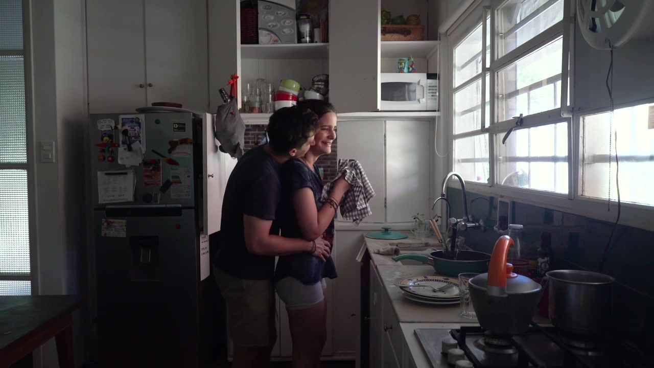 Namorado surpreende a namorada com um abraço na cozinha, em plano de perfil.