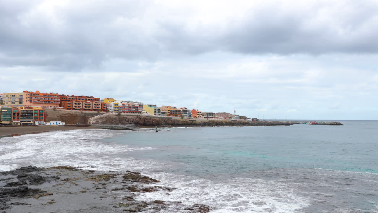 Calm sea moving slowly towards the peninsula with views of part of town lying on cliff with black beach and the statue of Neptune Grand Canary island valley 4k slow motion capture at 60fps