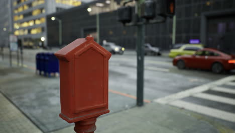 red mailbox stands prominently on a busy urban street during a drizzly evening
