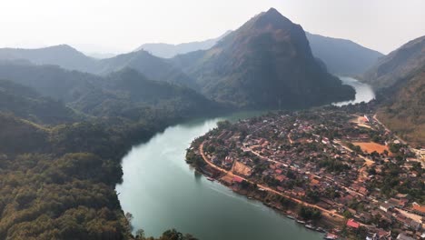 aerial view of nong khiaw, laos, where the nam ou river curves past a small riverside town nestled among lush green mountains and dramatic limestone peaks under a hazy sky