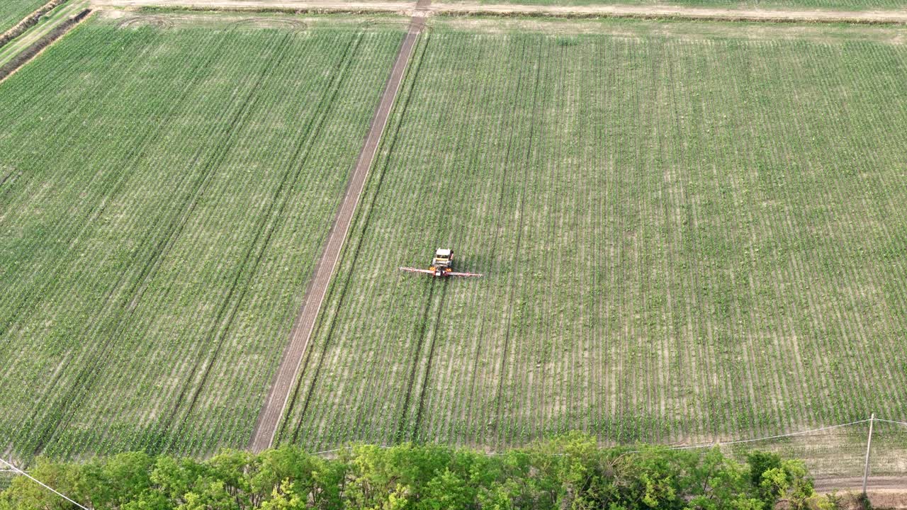 Aerial shot of a tractor with boom sprayer applying pesticides to a large monoculture maize field adjacent to a narrow tree line, illustrating environmental risks from intensive GMO crop management