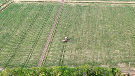 aerial shot of a tractor with boom sprayer applying pesticides to a large monoculture maize field adjacent to a narrow tree line, illustrating environmental risks from intensive gmo crop management