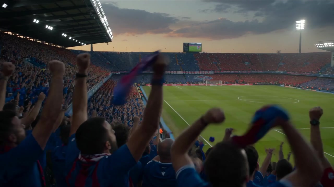 Aficionados locales animando, agitando bufandas y aplaudiendo en el estadio al anochecer, con focos y pantalla gigante.