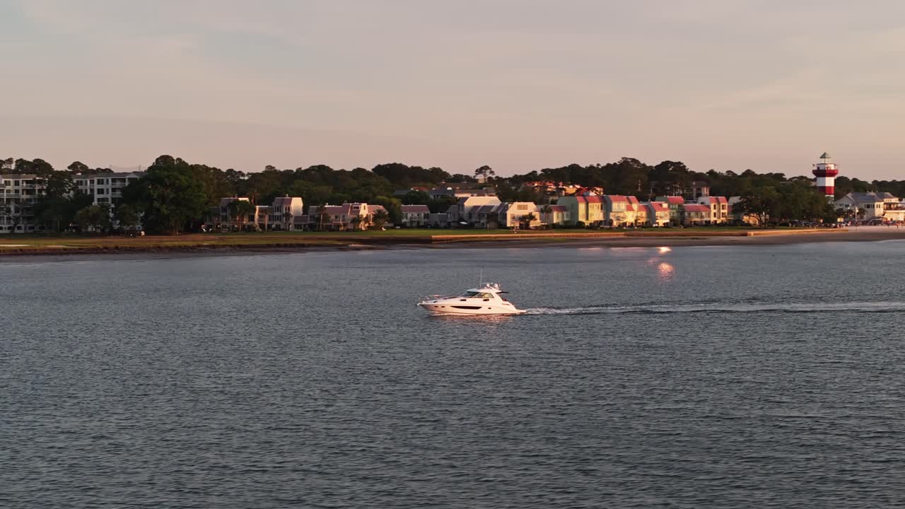 Drone tracking left follows yacht along coast passing over pier as golden sun fades, casting glow across open water surface, Harbour Town Pier South Carolina USA