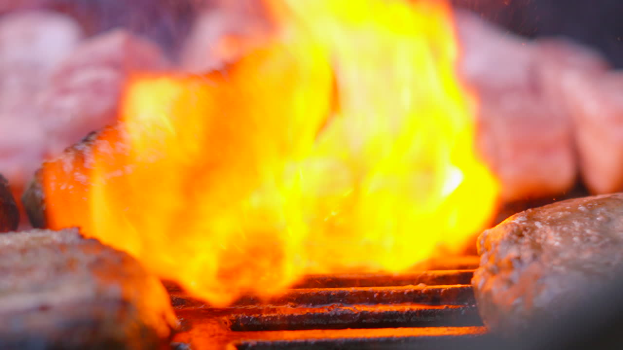 Close Up Slow Motion of Beef Burger Being Flipped and Tumbling on Hot BBQ Grill with Big Fire Flames Erupting from Below. High Quality Barbecue Summer Cooking Food. Pork Belly Slices in Background.