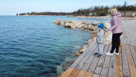 A-static-shot-of-a-mother-son-duo-standing-on-a-wooden-bridge-along-a-sea-shore-with-the-son-trying-to-throw-rocks-in-the-sea-and-the-mother-guarding-him