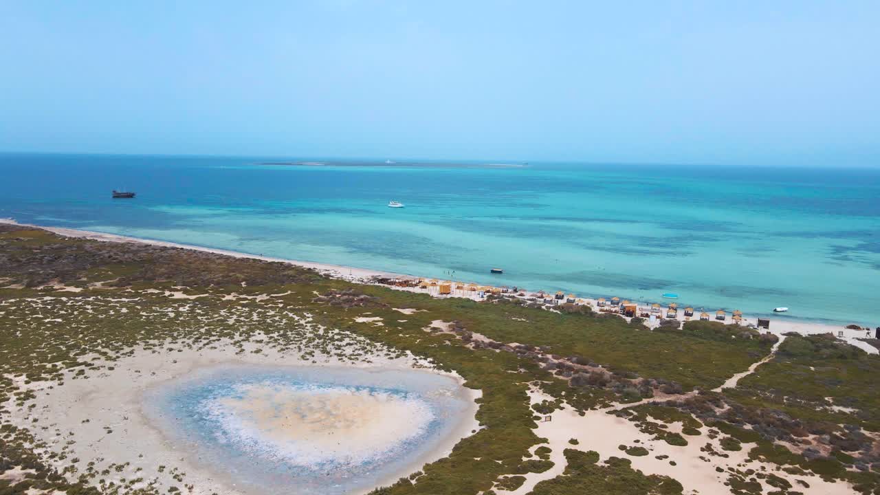 A blue ocean with boats and people enjoying the day. The beach is deserted with just a few people scattered around. There are small white rocks along the shore, which adds to the beautiful landscape.