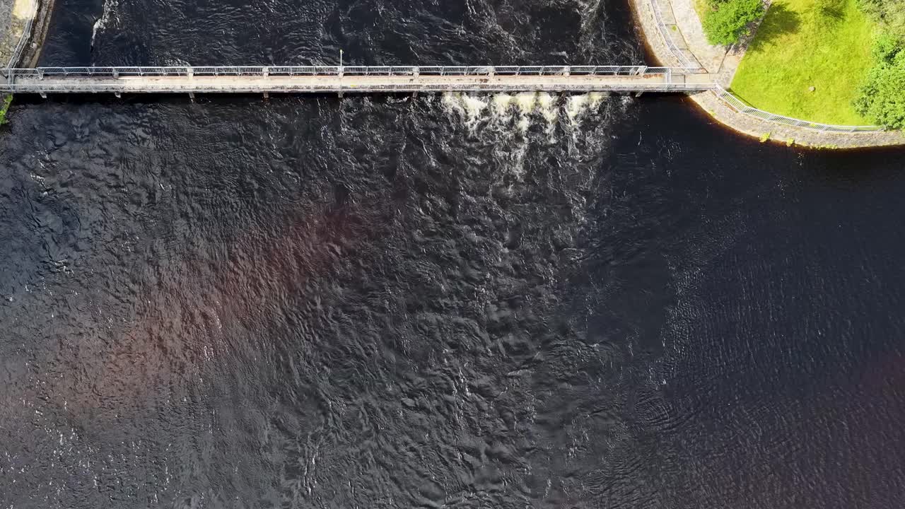 Drone captures hydroelectric dam, water flow, and green landscape in bright daylight, smooth overhead movement