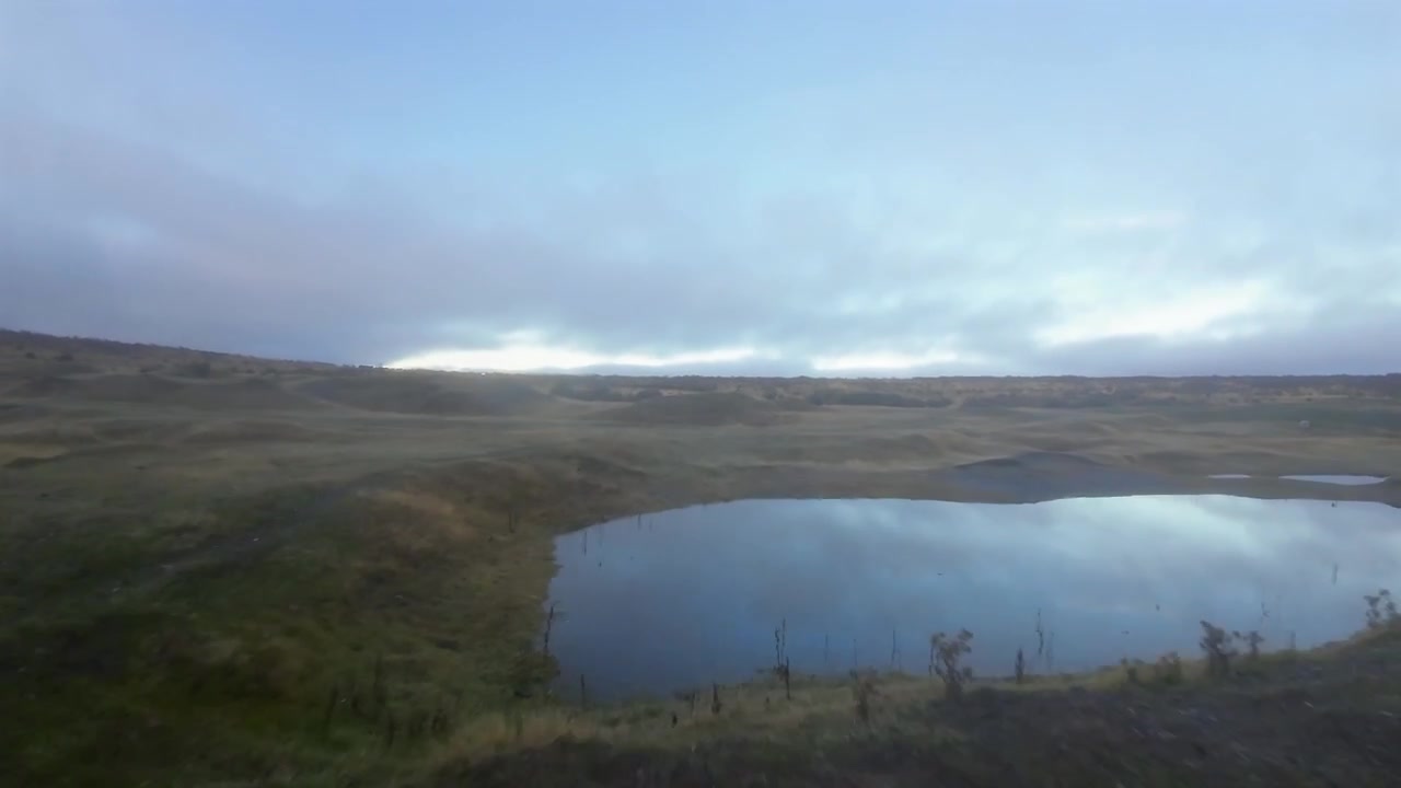 FPV aerial descending toward a small mirror still lagoon cradled in golden steppe, revealing the quiet vastness of southern Patagonia