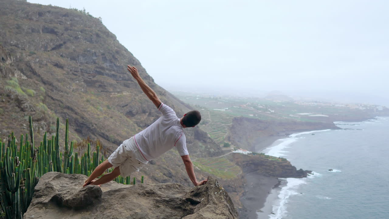 Standing on one hand in the mountains, the man's back to the camera, he focuses on the ocean view, engrossed in meditation