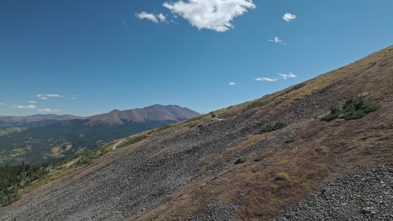 Drone dolly to evergreen high alpine mountain side with car at center driving along dirt road