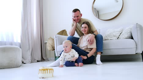 beautiful modern caucasian family of three at home. little baby sitting on the floor with a cake in front of him. low angle view.