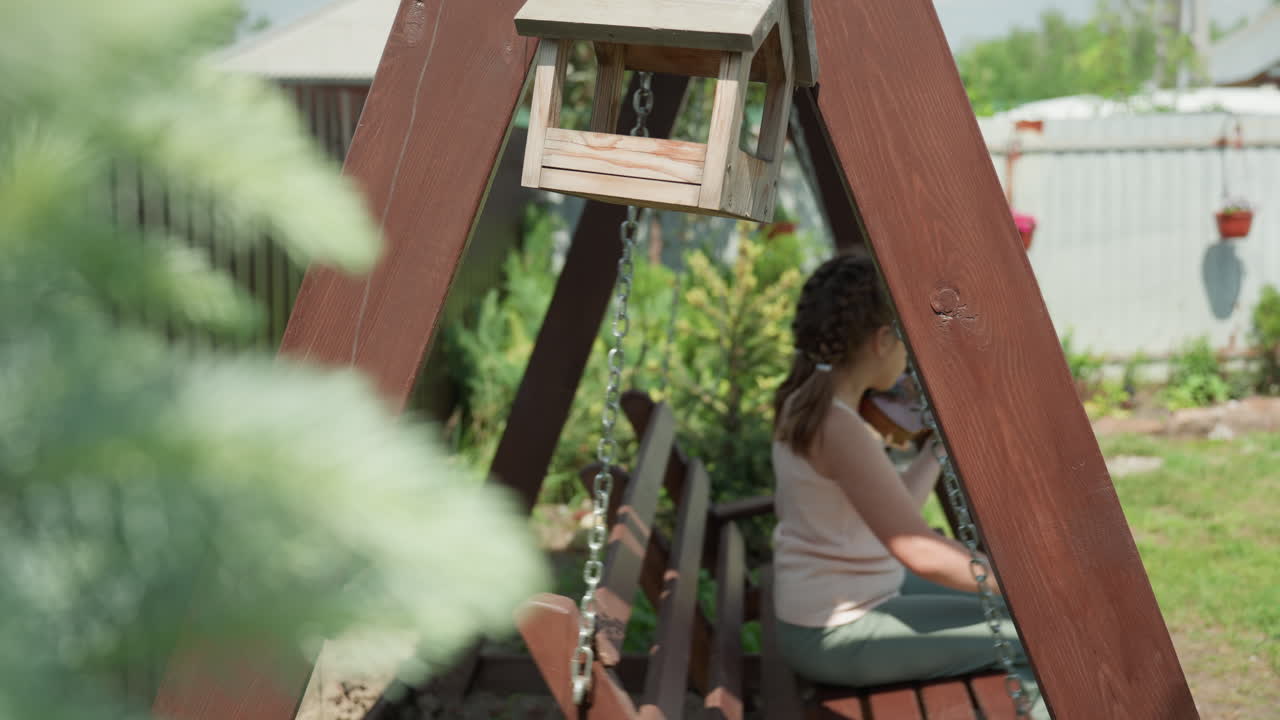 Young Girl Outdoors With Violin, Girl With Bow In Sunlit Garden Playing Violin On Swing, Casual Summergear Girl Attentively Practicing Violin Outdoors Amidst Lush Greenery And Fencing