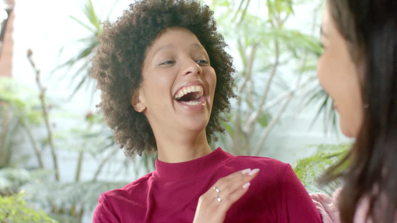 Madre e hija riendo tras un comentario; la madre señala y toca el brazo en medio de plantas verdes
