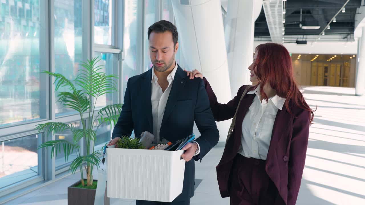 Sad European man walking with personal belongings in white box while Latina colleague comforts him in modern office corridor after termination or resignation
