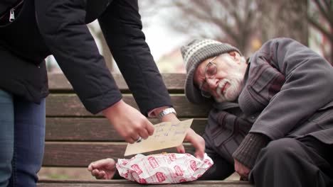 a caring stranger offers a warm meal to an elderly man sitting on a bench in a park. the winter atmosphere highlights the importance of compassion and generosity in tough times.