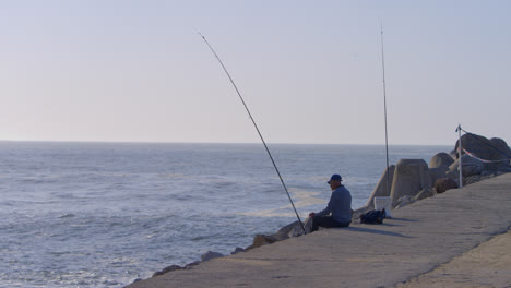 Man-fishing-on-the-harbor-break-wall-in-the-town-of-Nazare,-Portugal