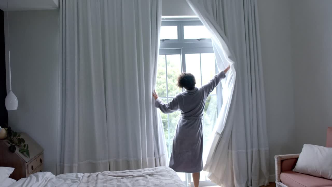 Schiena di donna birazziale in accappatoio che apre le tende in camera da letto, al rallentatore, spazio per il testo