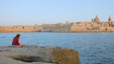 Mujer-Leyendo-Un-Libro-Sentada-Sobre-La-Costa-Rocosa-Del-Mar-Con-Vistas-A-La-Ciudad-De-Valletta-En-Malta