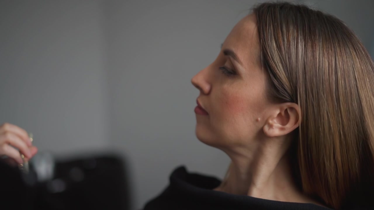 Primo piano di una donna che si sistema i capelli con movimenti delle mani davanti a uno sfondo grigio uniforme. I capelli sono lisci e castano chiaro, il soggetto indossa un top nero. L'illuminazione soffusa crea un'illuminazione uniforme.
