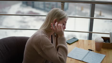 la joven escribe pensativa, hace una pausa para poner la mano en la mandíbula, continúa escribiendo con el teléfono y el café en la mesa, el fondo presenta una escena de invierno borrosa a través de la ventana